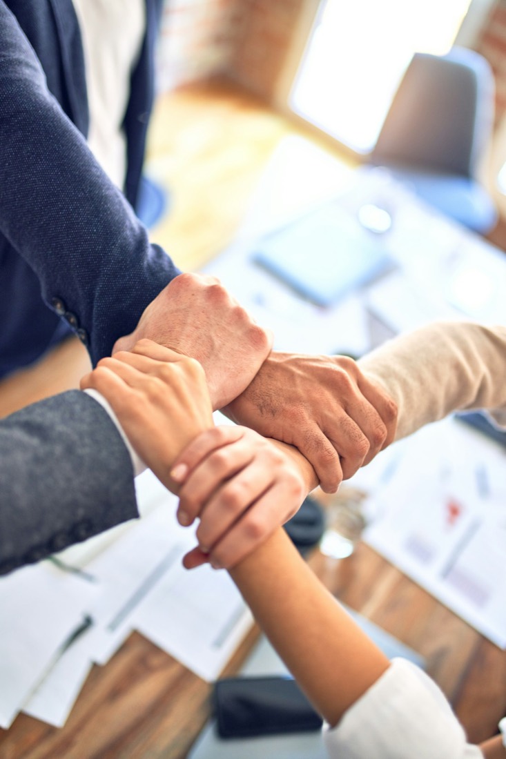 Colleagues joining hands over a desk, symbolizing teams coming together